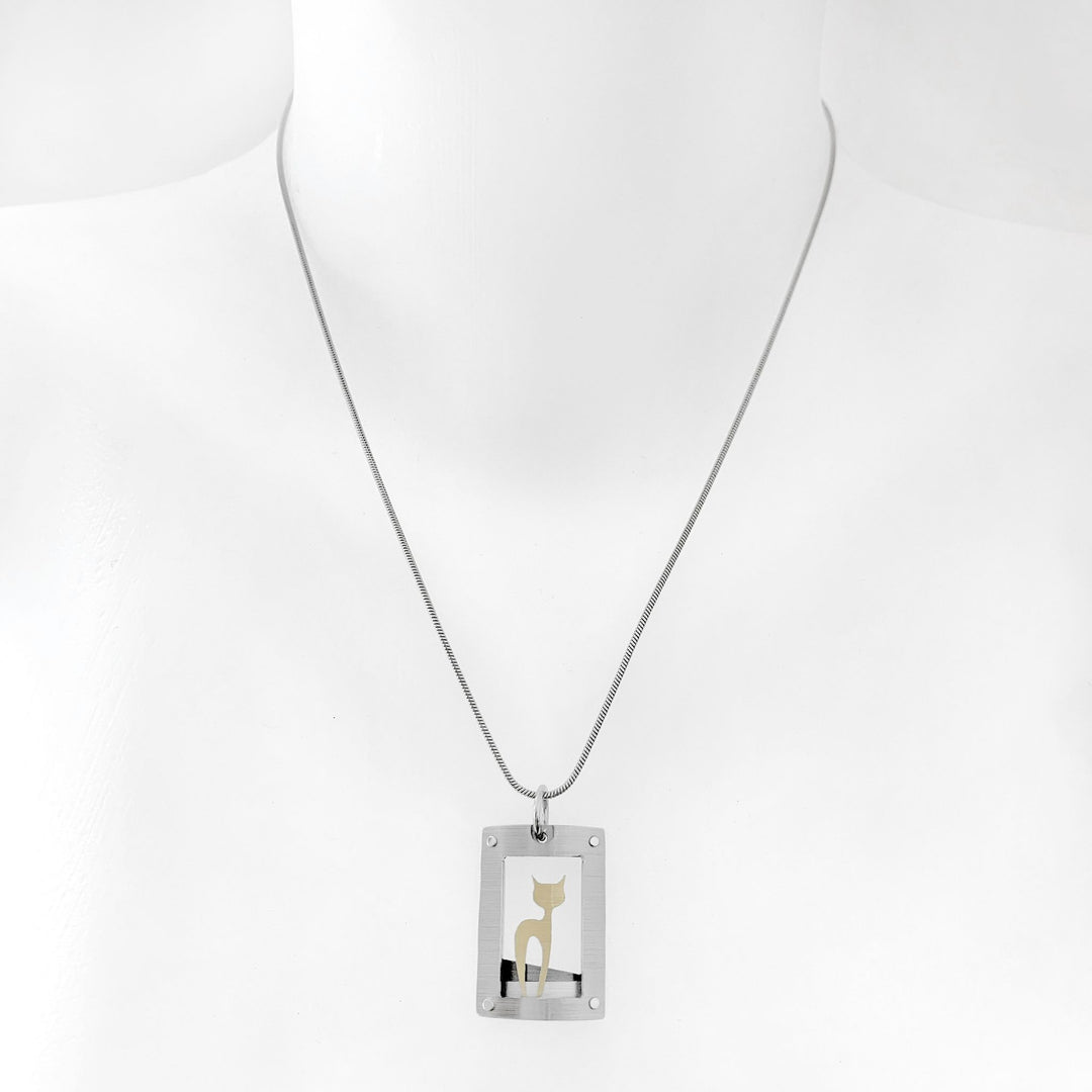 This cat necklace with pendant has a curved, grey frame with gold-colour cat silhouette inside. Side-view of cat with head turned to side so both ears are visible. Minimalist design. By JR Franco. Stainless steel snake chain included.