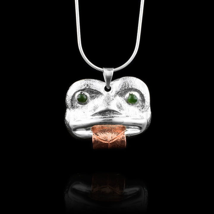 This Frog pendant depicts the face of the Frog which is done in sterling silver and has a copper band extending from the piece depicting its tongue. The eyes of the Frog are made from genuine BC jade. There are details carved onto the tongue and the face has been shaped with embossing.