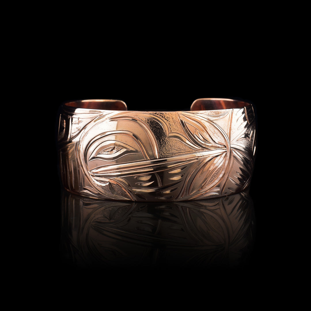 This copper cuff bracelet has a single, wide band with a gap in the back. The baby Hummingbird is depicted in the center with its head surrounded by an oval designs representing the womb. Where its beak meets the womb on the right, there are petals designs. The head of the mother Hummingbird is carved on the right side and the left depicts her outstretched wing.