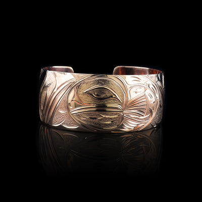 This copper cuff bracelet has a single, wide band with a gap in the back. There is a depiction of the Baby Salmon in its egg carved into the band. In the egg, you can see the eye of the baby Salmon and a fin sticking out on the right side. There are two Salmons depicted on either end of the band watching over the egg and there are forest and river details carved behind them behind them.