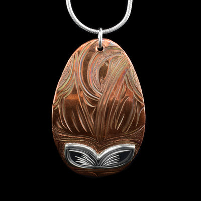 This Hummingbird pendant is made from sterling silver nad copper with a large, oval shape. There are depcitons of the Hummingbird and a flower carved into the surface of the piece. The depiction of the flower has been made using silver.