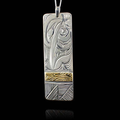 This Hummingbird pendant is made from sterling silver and 14K gold with a long, rectangular shape. There are carvings on the surface of the pendant depicting the Hummingbird and floral designs. There is a 14K gold strip 3/4's of the way down the piece.