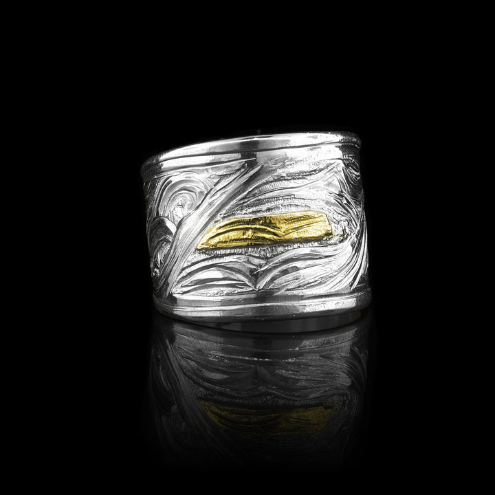 This Orca ring has a wide sterling silver band that has wavy designs and carvings made across the surface of the band. There is a depiction of the Orca made from 14K gold that is carved into the band. The Orca is a small rectangle with details made in it. 