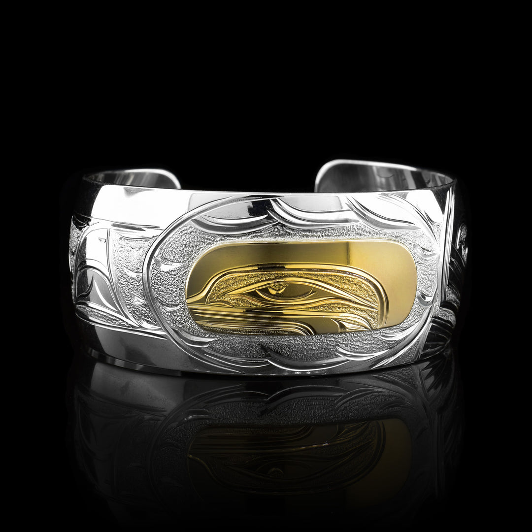 This stunning Orca bracelet is made using sterling silver, 14K gold and has a single, wide band with a gap in the back. There are intricate carvings on the surface of the piece that depicts the Baby Orca, done in gold, in the womb and being watched over by its mother who is depicted in silver and carved on the side of the piece. On the other side of the band, there are depictions dorsal fins in the sea from the Orca pod.