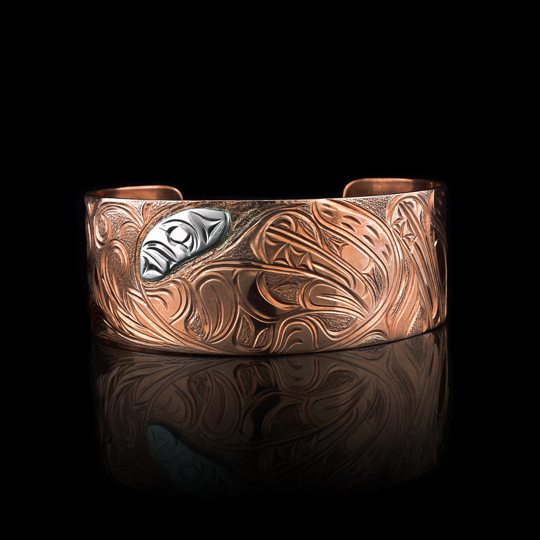 This Bear bracelet has a wide band made from copper and with a gap in the back. In the center, the baby Bear is depicted with its head facing left and its paws curlced up in front of it. Around the baby there is an oval designs which represents the womb and there is a sterling silver oval with carvings on it to the opposite side of the baby. On the right end of the band, the mother bear is depicted with a side profile of her head which is tilted upwards. On the other side, there are floral designs