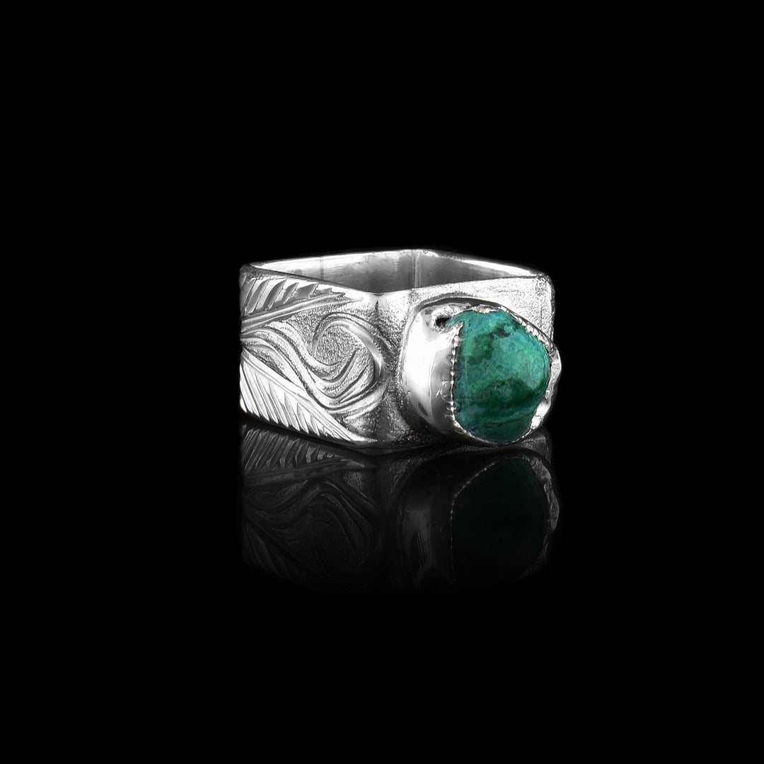 This aquamarine silver ring has a heavy gauge and wide band. The band also has a square shape and there are swirling carvings made across the band depicting flowers. There is a roughly cut aquamarine gem set on top of the ring.