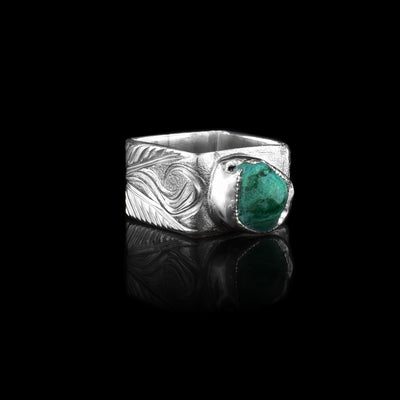 This aquamarine silver ring has a heavy gauge and wide band. The band also has a square shape and there are swirling carvings made across the band depicting flowers. There is a roughly cut aquamarine gem set on top of the ring.