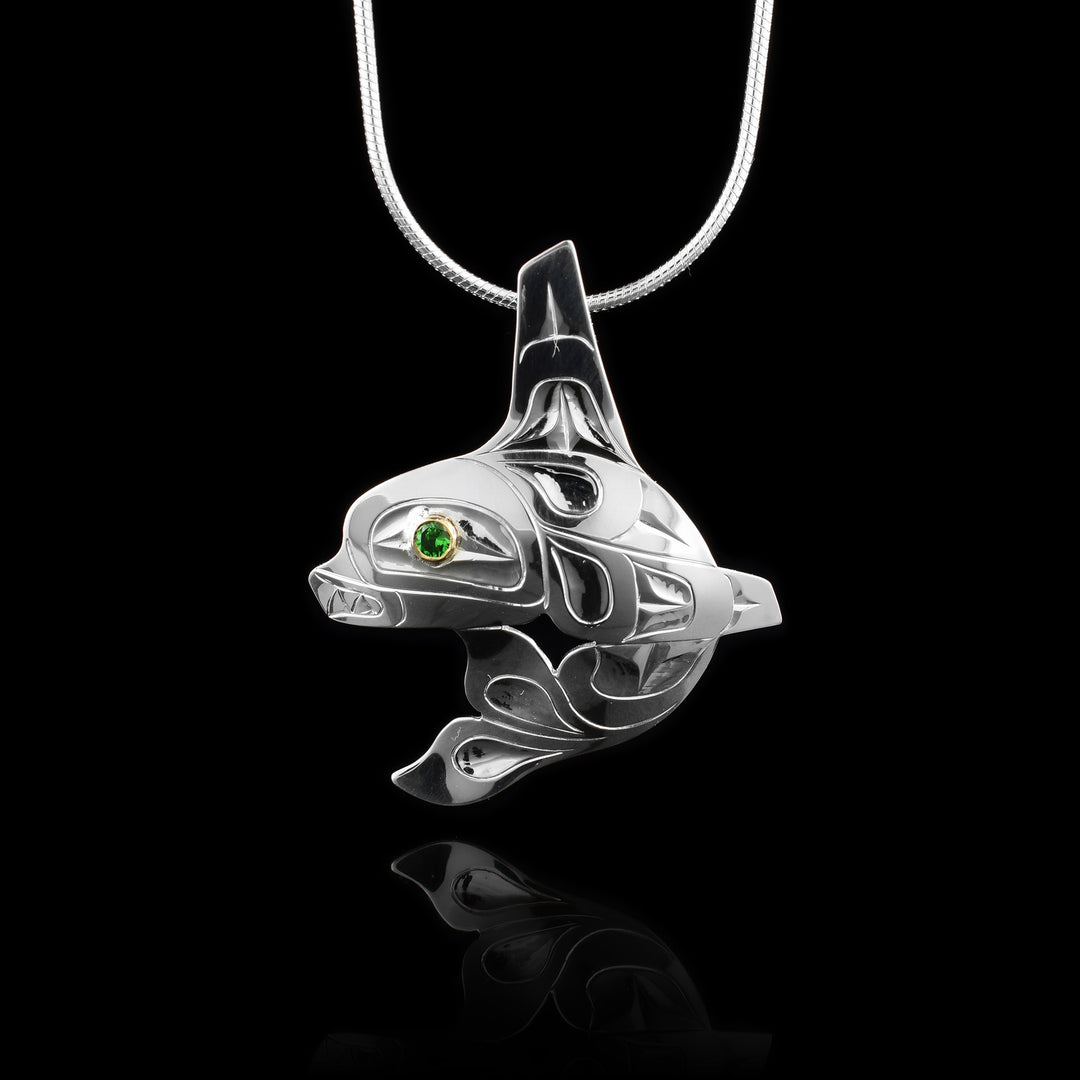 This Orca pendant is made from sterling silver and takes the shape of the Legend. The Orca is at a side profile with its tail curled underneath it and it has a large, dorsal fin extending from the top of it. There is another fin at its side and carvings across its surface depicting the eye, teeth as well as tail and fin details. There is a green tsavorite gem embossed in 14K gold in the eye of the Orca.