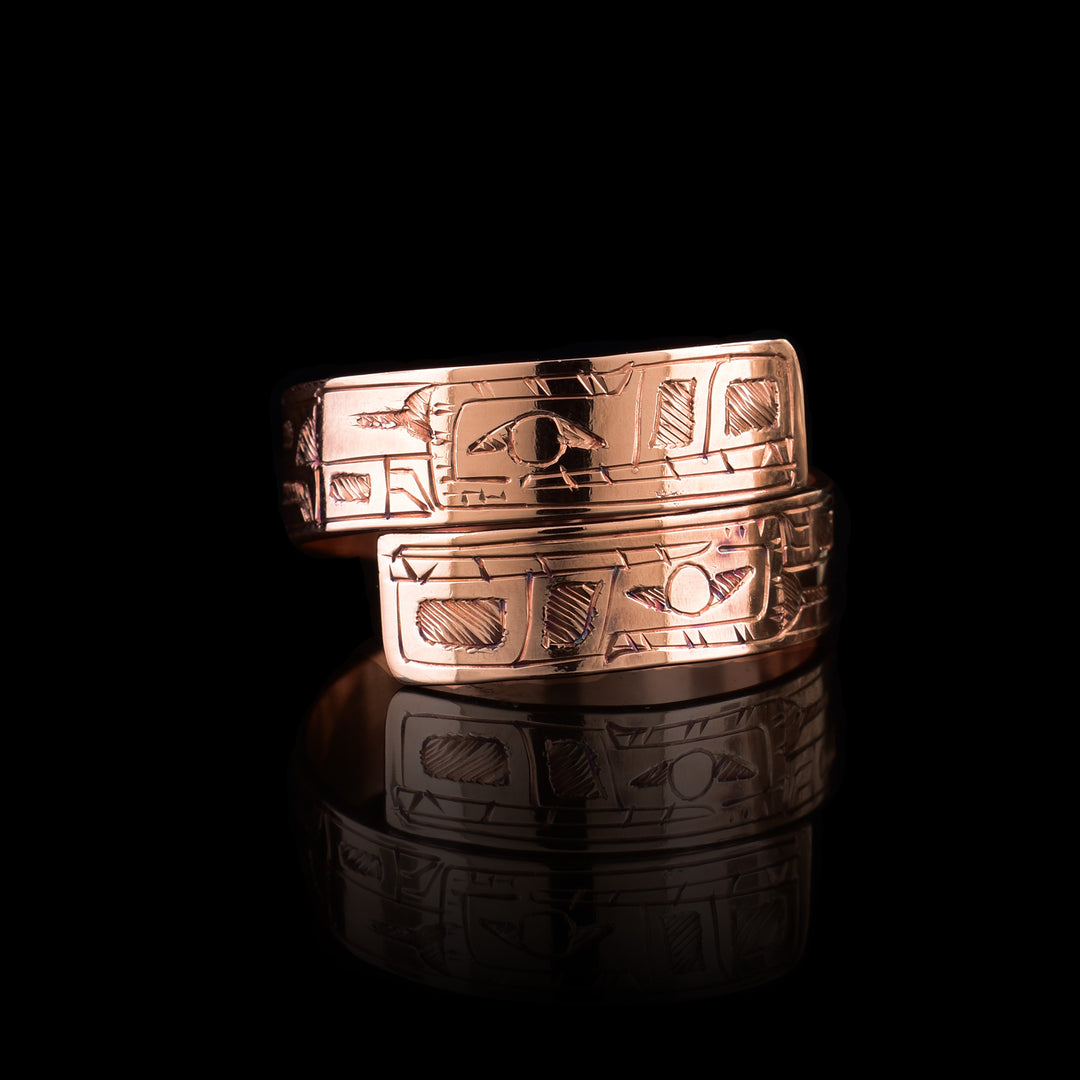This copper wrap around ring has a single band that wraps around the finger twice. There is a depiction of the Bear head at one end of the band and another on the other end. The depictions are done in a side profile with hatching design in the background of the scene.