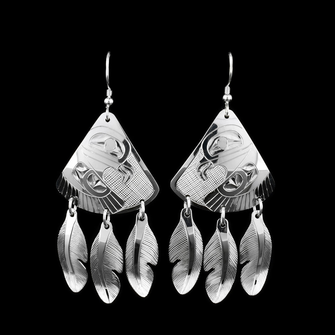 These Eagle feather earrings have sterling silver hangs that are wide and teardrop shaped. The Eagle is shown at a side profile with its wing below its head. There are feather details and the wing and cross hatching deisgns in the background of the piece. There are three, sterling silver feathers dangling from jump rings below the hangs.