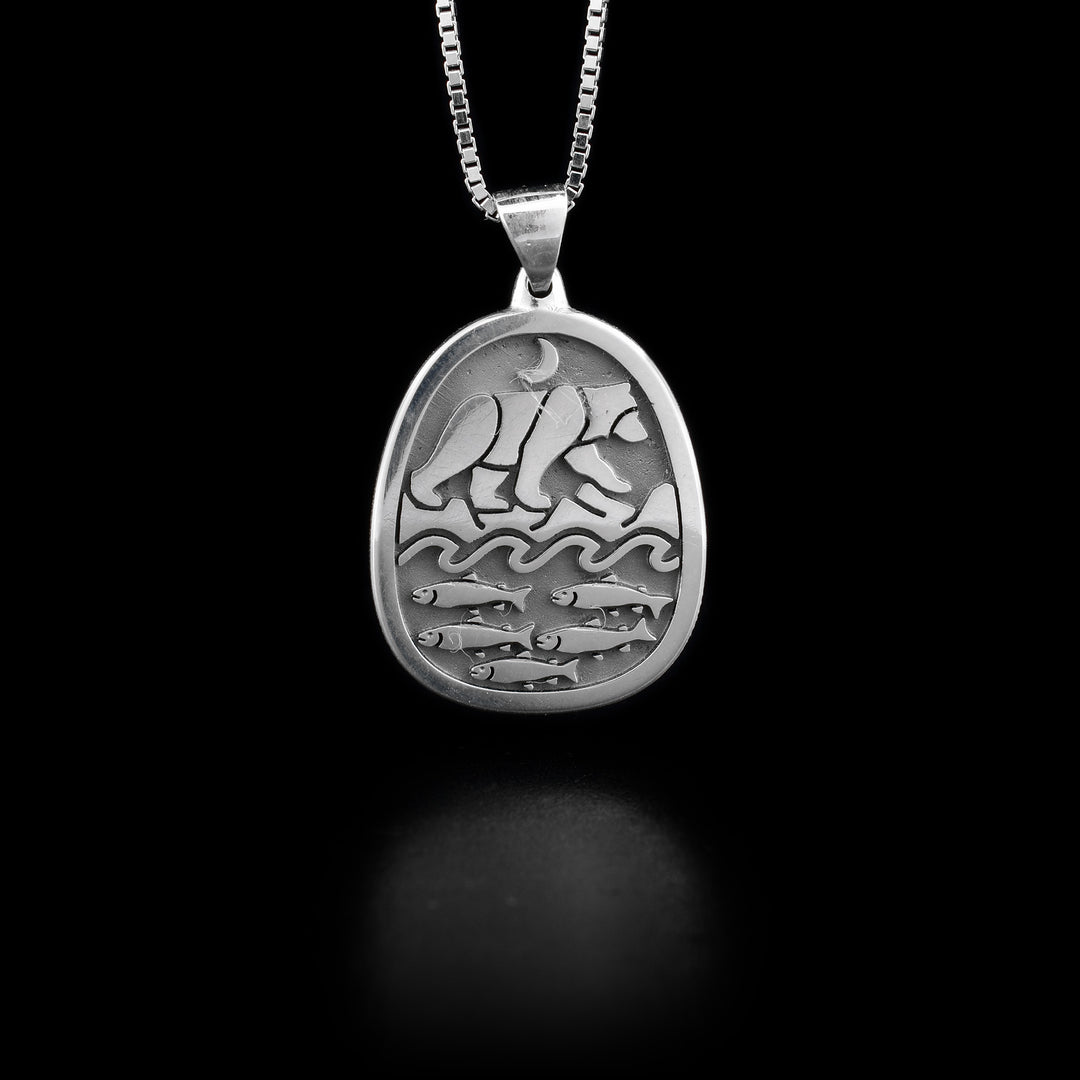 A silver, oval-shaped salmon and bear pendant with a wide bail. It depicts a bear walking along a river full of salmon. There is a moon above the bear.