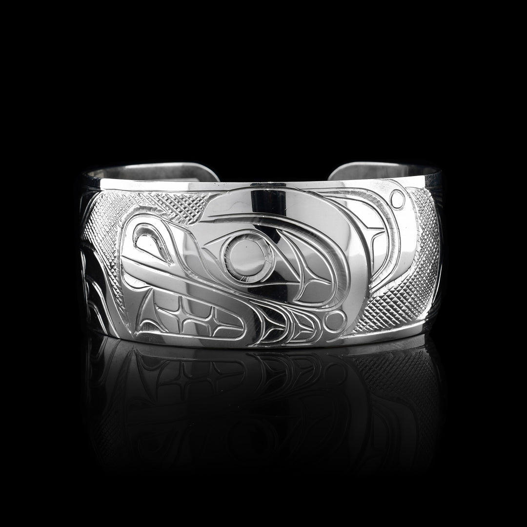 This Bear bracelet has a wide, sterling silver band with a gap in the back. The Bear's head is carved in a side profile in the center of the band and takes up the entire width of the band. The head is facing the left and in front of it, its clawed paw is shown. There are cross hatching patterns in the background of this piece.