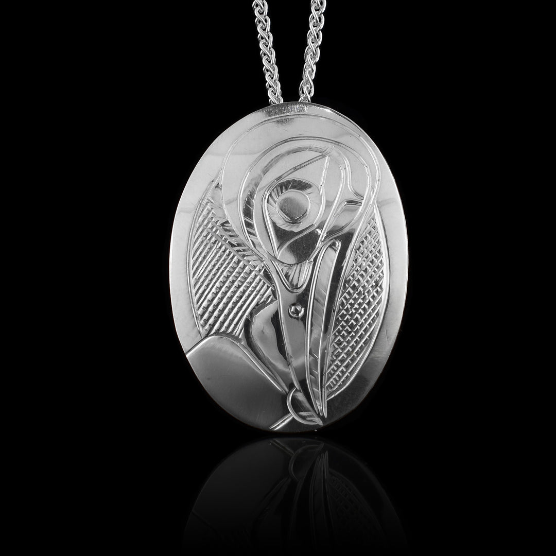 This Hummingbird pendant has been made form sterling silver and has a wide, oval shape that is longer than it is wide. The head of the Hummingbird is carved at a side profile looking downwards from the piece. Its beak extends to the bottom center and there are cross hatching patterns in the background.
