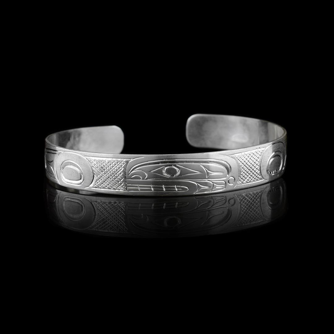 This dainty Orca bracelet has a thin, sterling silver band with a gap in the back. The head of the Orca is carved in the center of the band and takes up the width of the band. The head has an oval shape, is at a side profile and its facing the left. On both ends, there are circular, oblong and triangular patterns depicting the rest of the legend. There are cross hatching designs in the background of this piece.