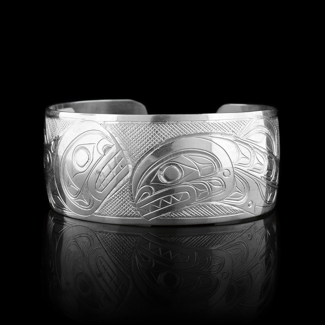 This Salmon and Orca bracelet has a wide, sterling silver band with a gap in the back. The body of the entire Salmon is depicted in a side profile on the left of the piece. This legend has a signature hump on its back and its head is facing upwards in the center of the piece. The entire Orca is depicted on the right. It has its dorsal fin and its head is breaching downwards in the center of the band. There are cross hatching patterns in the background of the piece.