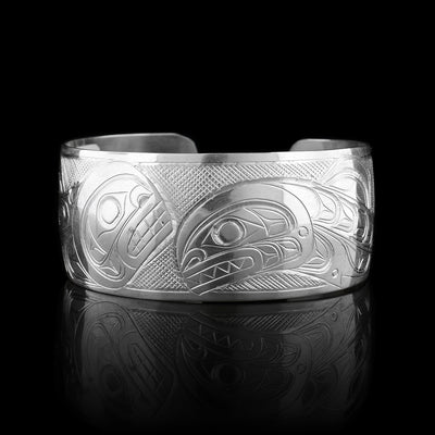 This Salmon and Orca bracelet has a wide, sterling silver band with a gap in the back. The body of the entire Salmon is depicted in a side profile on the left of the piece. This legend has a signature hump on its back and its head is facing upwards in the center of the piece. The entire Orca is depicted on the right. It has its dorsal fin and its head is breaching downwards in the center of the band. There are cross hatching patterns in the background of the piece.