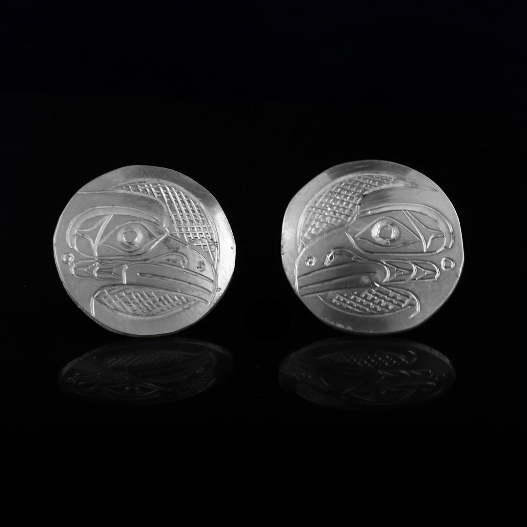 These Raven stud earrings are made from sterling silver and are circle shaped. There are carvings of the side profile of the head of the Raven legend made into the surface of the pieces. In the space behind the Raven head, there are cross hatching designs.