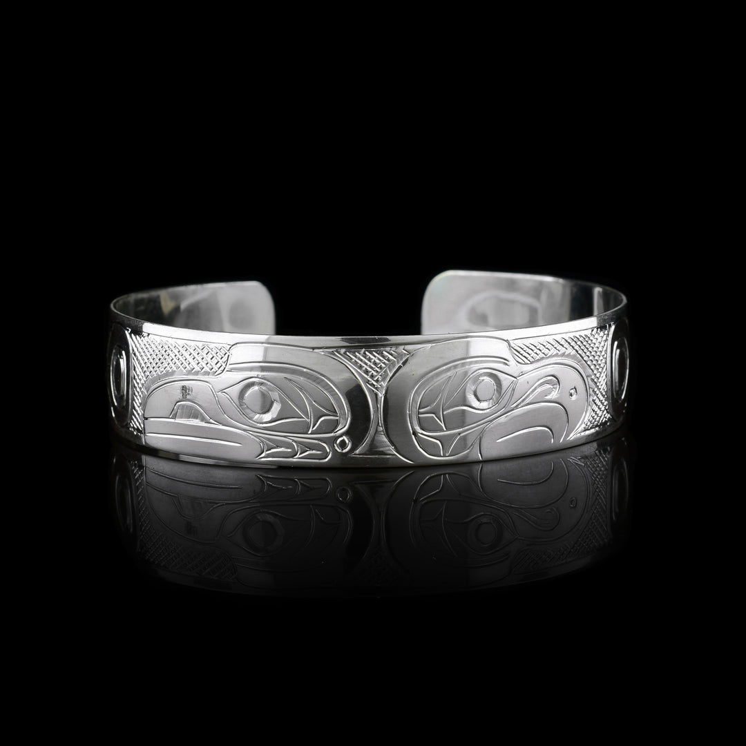 This sterling silver cuff bracelet has a single band with a gap in the back. The heads of the Raven and Eagle legend are carved into the center of the piece and are facing away from eachother. The heads take up the width of the band with the Raven on the left and the Eagle on the Right. There are circular and triangular designs depicting their wings on both the ends of the piece.