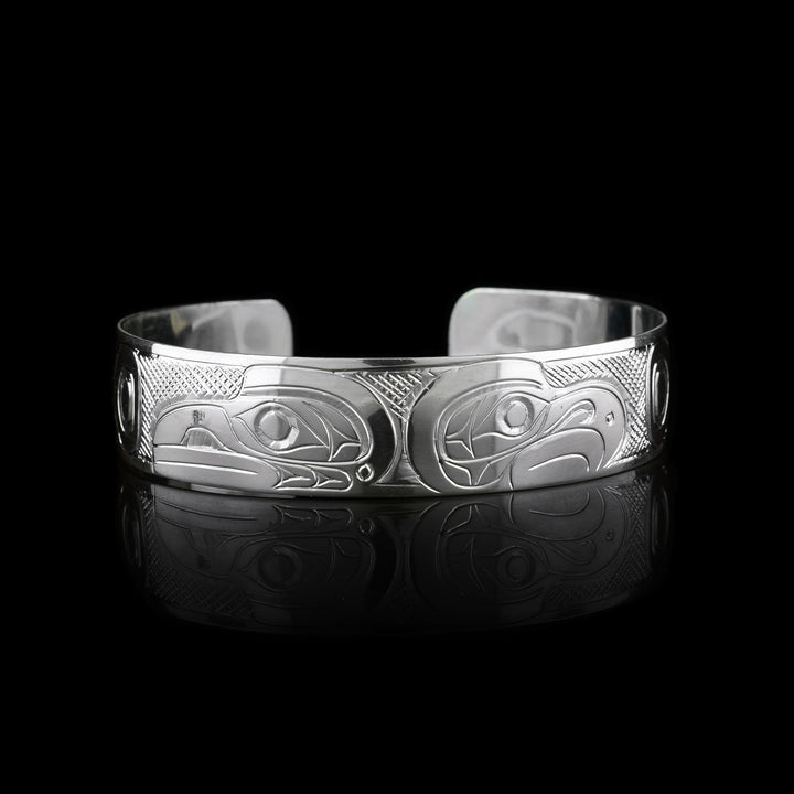This sterling silver cuff bracelet has a single band with a gap in the back. The heads of the Raven and Eagle legend are carved into the center of the piece and are facing away from eachother. The heads take up the width of the band with the Raven on the left and the Eagle on the Right. There are circular and triangular designs depicting their wings on both the ends of the piece.