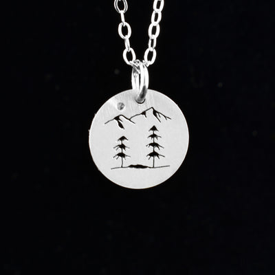 This trees and mountain pendant necklace has a simple chain with a small, sterling silver pendant dangling from a jump ring. The pendant is circle shaped and there are two trees with two mountain peaks above the trees. The designs are cutout into the pendant.