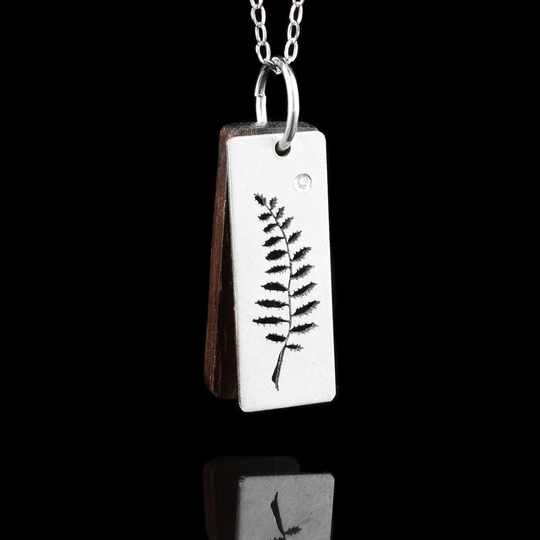 This fern leaf pendant has a sterling silver rectangle pendant with a cutout leaf pattern on it. The stem is long and the leaves have sharp edges on them. There is a dark, brown wooden piece behind the pendant that matches it in size.