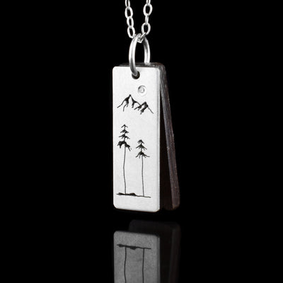 This mountain pendant has a sterling silver pendant that is thin and rectangular. There is a dark brown piece behind the silver pendant that has the same shape. There is a design with two mountains and two trees below the mountains. These deigns are etched into the silver.