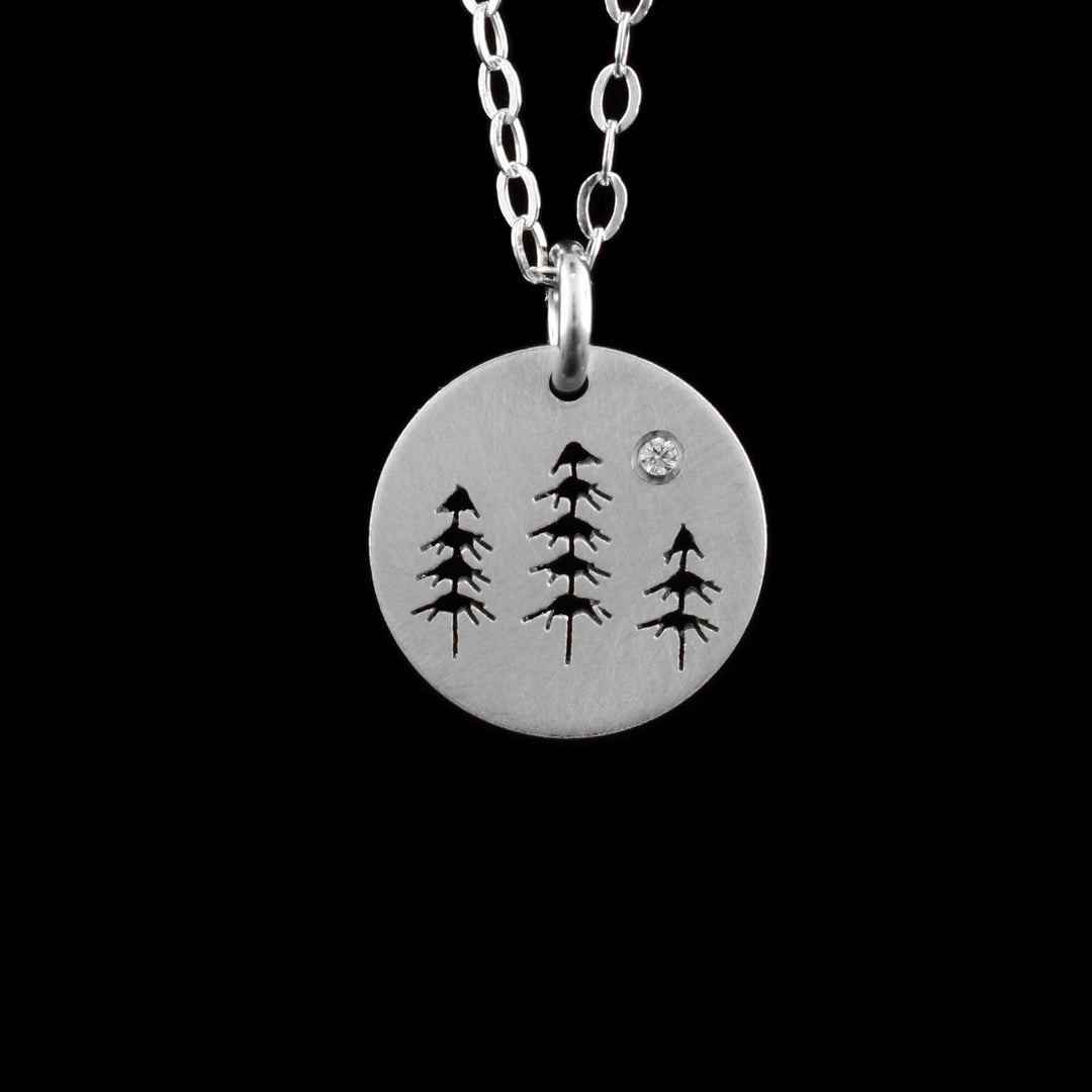 This trees and mountain pendant necklace has a simple chain with a small, sterling silver pendant dangling from a jump ring. The pendant is circle shaped and there are three trees with clumps of leaves going up them. The center one is in the center of the piece. The designs are cutout into the pendant.