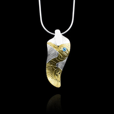 This feather gold pendant is made from sterling silver and 14K gold plating. The piece is feather shaped and there is a depiction of the Heron done in gold and  the feather is done in silver. The Heron is shown from the side with its head facing slightly upward. There are carvings on the surface of the piece depicting the Heron and feather details. There is a blue topaz gem embedded into the eye of the Heron.