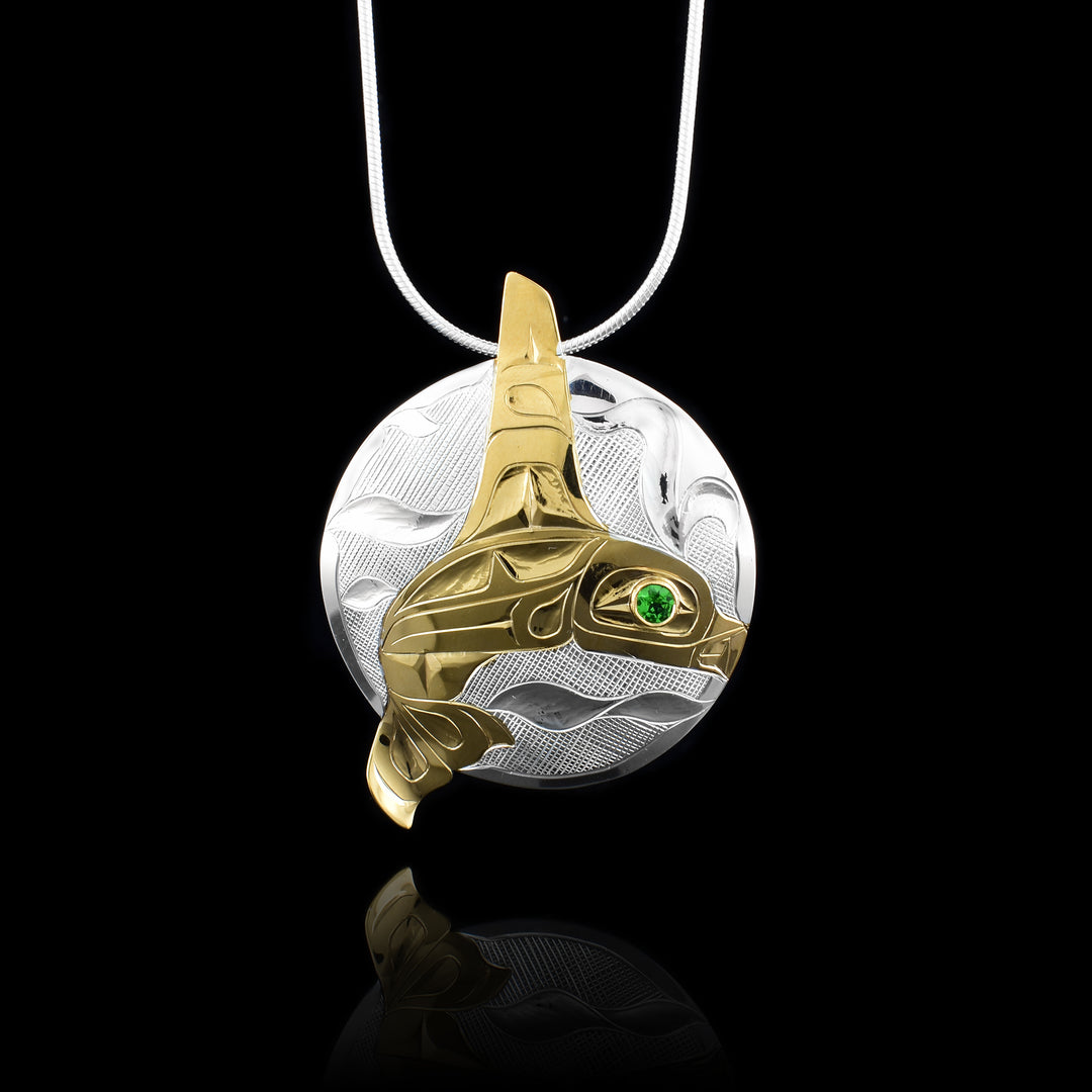 This Orca whale pendant is made from a sterling silver circle piece. The Orca legend is depicted in 14K gold plating and its dorsal fin extends past the circle at the top of the piece and its tail extends past at the bottom. There are carvings on the surface of the piece depict the details of the Orca in the ocean. On the silver circle, there are swirling designs depicting waves carved into it. There is a green tsavorite gem embedded in the eye of the legend.