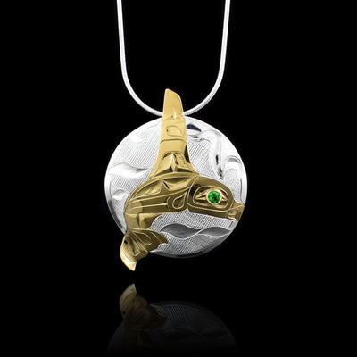 This Orca whale pendant is made from a sterling silver circle piece. The Orca legend is depicted in 14K gold plating and its dorsal fin extends past the circle at the top of the piece and its tail extends past at the bottom. There are carvings on the surface of the piece depict the details of the Orca in the ocean. On the silver circle, there are swirling designs depicting waves carved into it. There is a green tsavorite gem embedded in the eye of the legend.
