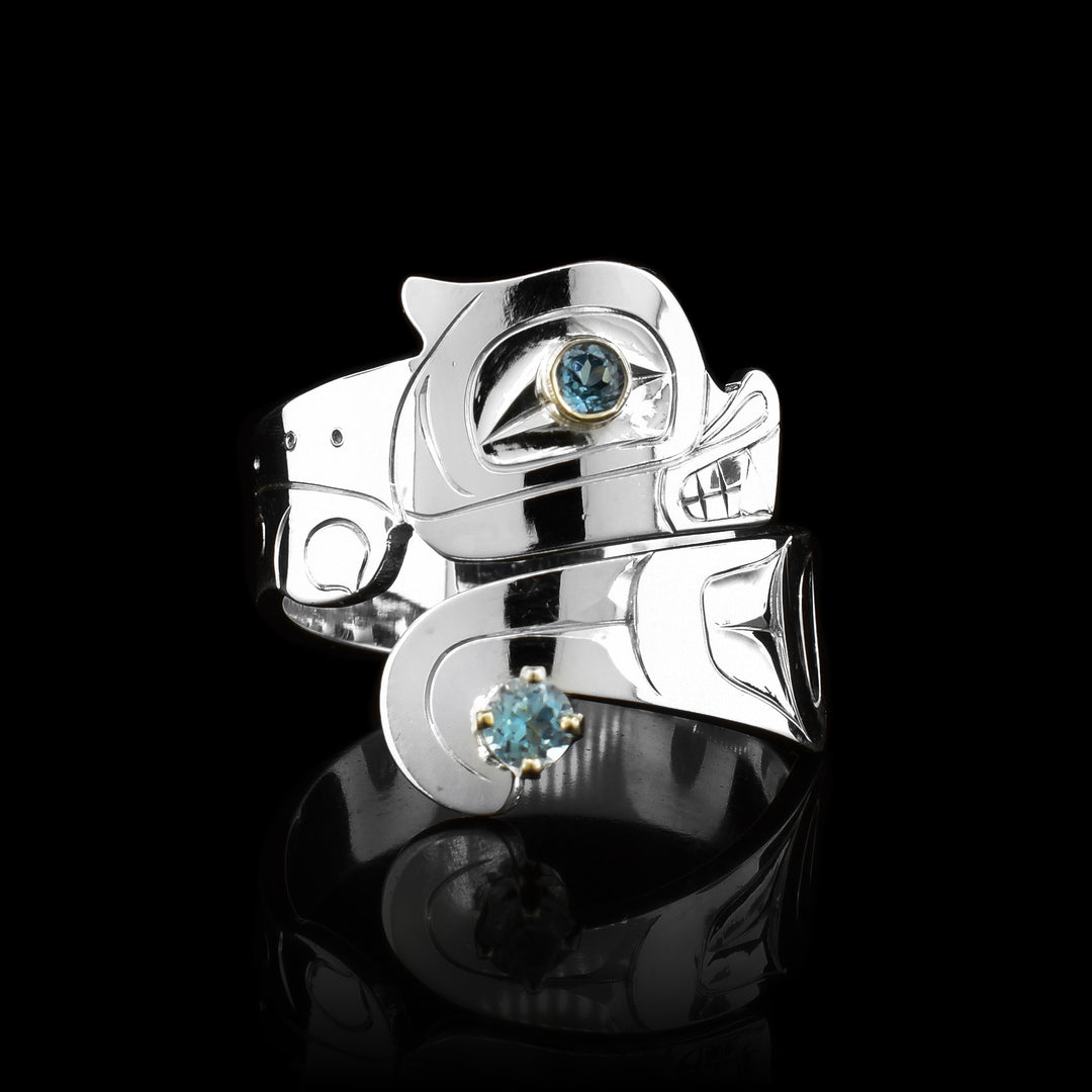 This Otter sterling silver blue topaz ring has a wide band that wraps around the finger twice. The top band is carved to depict the head of the Otter and the bottom one depicts its tail. There are blue topaz gems that are embedded into the eye of the legend and in the center of its tail. There are 14K gold highlights around the eye of the Otter.