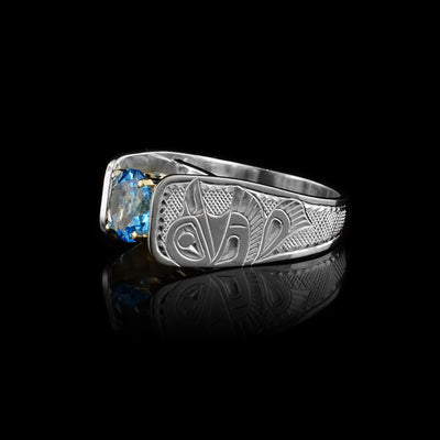 Solitaire Raven Ring with blue topaz by Fred Myra. The artist has hand-carved the profile of two Raven bodies on either side of the blue topaz he has set in the center of the ring. The background of the ring has been hand-carved into a neat crisscross pattern to allow for the Ravens to stand out. The round blue topaz has been set using a 14k gold claw setting. The ring tapers down at the back.
