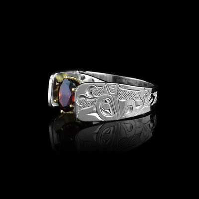 This Orca ring has a wide and tapered band that extends in cathedral style above the garnet gem that is set in 14K gold claws. There are carvings made on the band that depict the Orca. The Orca is facing the top of the ring with a water spout coming out of its head. There are cross hatching patterns in the background on the band.