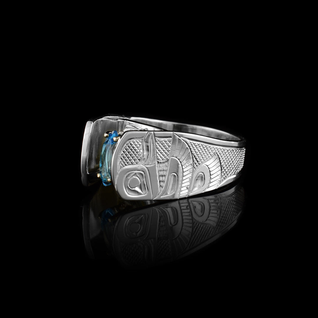 Solitaire Hummingbird Ring with blue topaz by Fred Myra. The artist has hand-carved the profile of two hummingbird bodies on either side of the blue topaz he has set in the center of the ring. The background of the ring has been hand-carved into a neat crisscross pattern to allow for the hummingbirds to stand out. The blue topaz has been set using a 14k gold claw setting. The ring tapers down at the back.