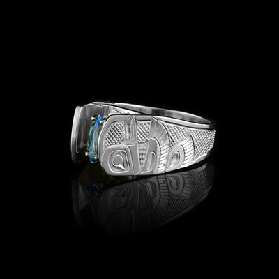 Solitaire Hummingbird Ring with blue topaz by Fred Myra. The artist has hand-carved the profile of two hummingbird bodies on either side of the blue topaz he has set in the center of the ring. The background of the ring has been hand-carved into a neat crisscross pattern to allow for the hummingbirds to stand out. The blue topaz has been set using a 14k gold claw setting. The ring tapers down at the back.