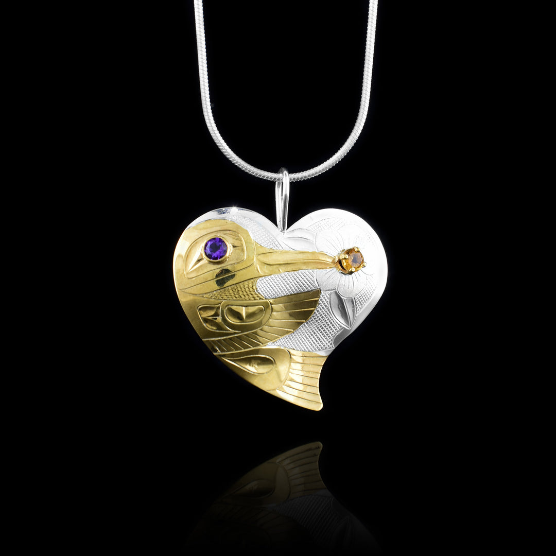 This citrine and amethyst pendant has a sterling silver hear shape with a Hummingbird depicted in 14K gold plated material on the side. There are carvings on the surface of the piece depicting the details of the Hummingbird who is shown feeding from a flower. The flower is done of the other end of the heart pendant and has leaf details around the petals. There is an amethyst in the eye of the legend and a citrine in the center of the flower.