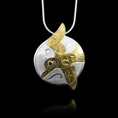 This Flying 14K Gold Hummingbird Pendant has a sterling silver circle piece. The Hummingbird legend is done in gold and is in flight. It has its wings outstretched and they extend past the circle portion of the piece at the top and the bottom. There are carvings on the surface depicting the Hummingbird with wind designs around it. There is a sapphire gem embedded in the eye.