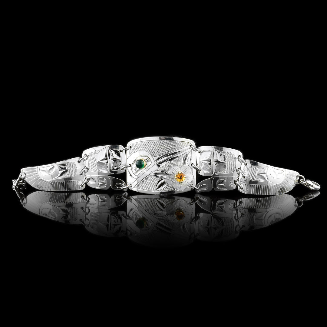 This beautiful panel bracelet has a sterling silver, rectangle panel with a depiction of the Hummingbird and a flower carved into it. There is a green onyx gem in the eye of the legend and a citrine gem in the center of the flower. There are two other panels on either side of the center one that taper. There are designs carved into these panels with the last ones depicting wings of the Hummingbird.