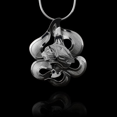 This Orca whale pendant is made from sterling silver and shows the Orca legend mounted on cutout pieces representing ocean waves. The Orca is in the center and has a large, dorsal fin with a tail curved in front of it. There are carvings on the piece depicting the scene.