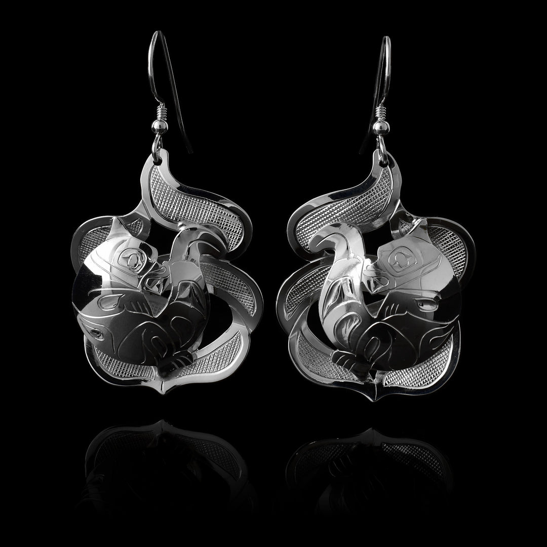 The Otter earrings are made using sterling silver and have abstract hangs depicting the Otter legend in ocean waves. The otter is curled up on its back with its tail above its head. The waves are made with cutout pieces and the Otter is set in the front of then. There are carvings made on the piece depicting the scene.