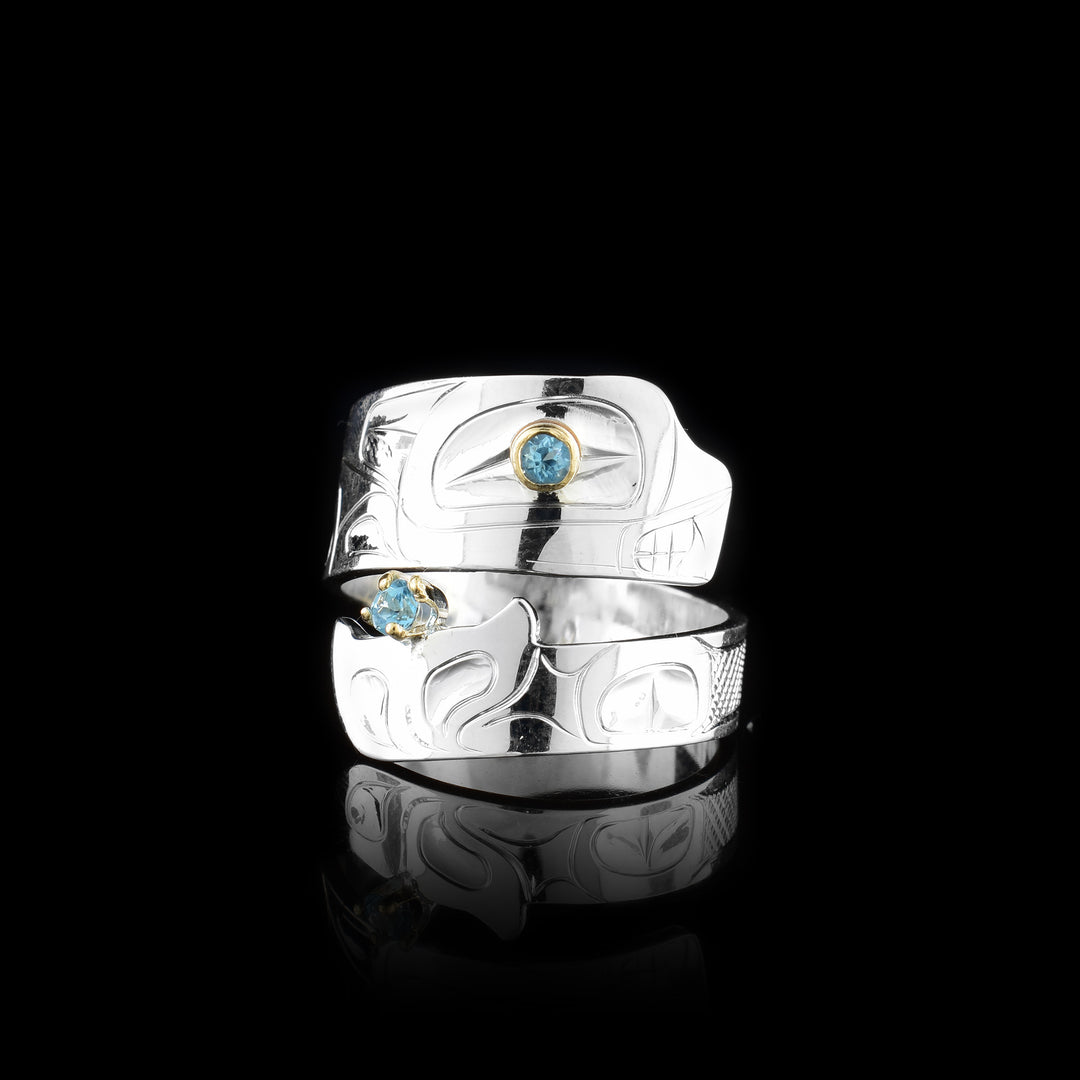 This blue topaz ring has a sterling silver band that wraps around the finger twice. The piece depicts the Orca and there are details of the legend carved into the band. The head is at the top with a blue topaz gem set in the eye and the bottom is the tail with another gem held in it.