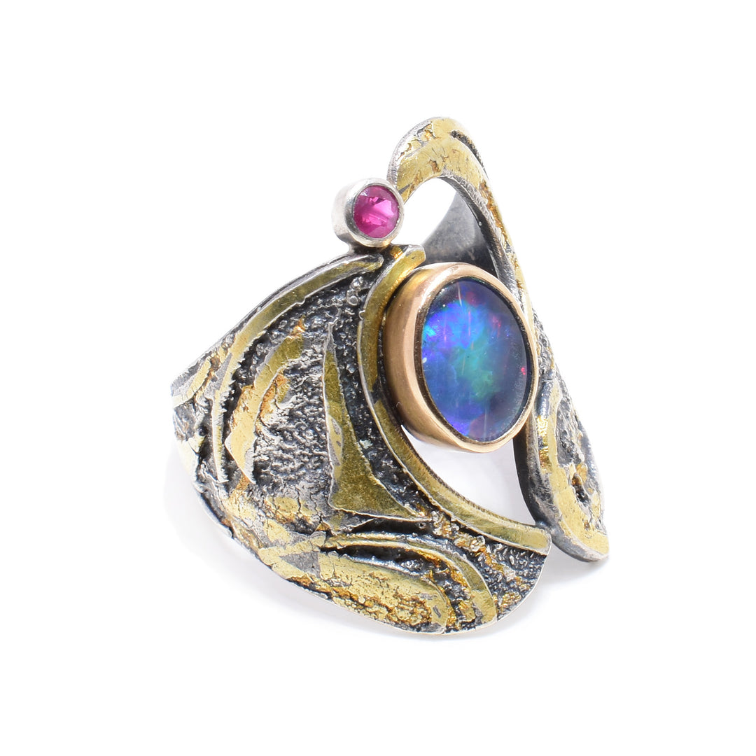 This unique opal ring is made from oxidized sterling silver. The silver has designs carved into it with 24K gold highlights. In the middle of the ring is an opal gem wrapped in 14K gold and a ruby wrapped in silver. 