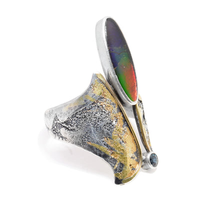 This unique sterling silver ammolite ring has 24K gold highlights and 14K gold bands in the middle of the ring. In the middle of the ring is a large, oval ammolite gem and a sapphire gem both wrapped in silver.