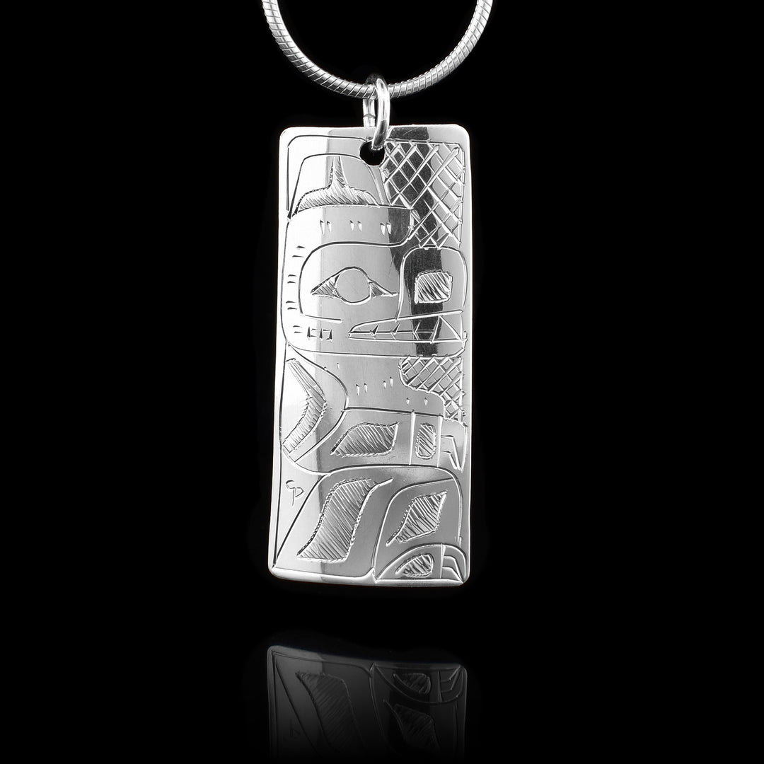 This Raven and Bear pendant is made from sterling silver and has a wide, rectangle shape. The bear is shown on this side at a side profile and it is sitting up right with its ears, eye, mouth and its front and back legs shown.