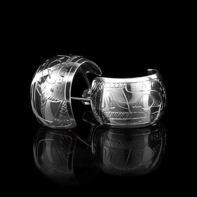 These wide hoop earrings are made from sterling silver and have a single, wide band with a gap in the back. There are carvings on the surface of the hoops depicting the Eagle.