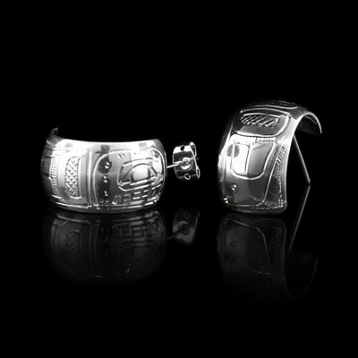 These wide hoop earrings are made from sterling silver and have a single, wide band with a gap in the back. There are carvings on the surface of the hoops depicting the Orca.