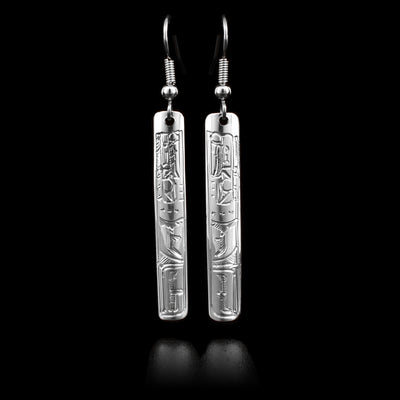 These sterling silver dangle earrings have long, thin, rectangular hangs with depictions of the Raven carved into them.