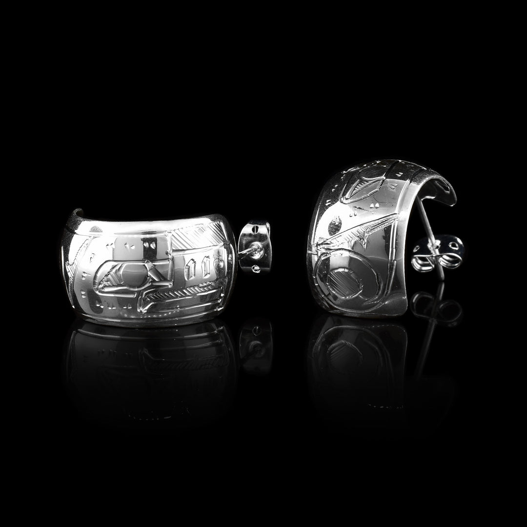 These wide hoop earrings are made from sterling silver and have a single, wide band with a gap in the back. There are carvings on the surface of the hoops depicting the Thunderbird.