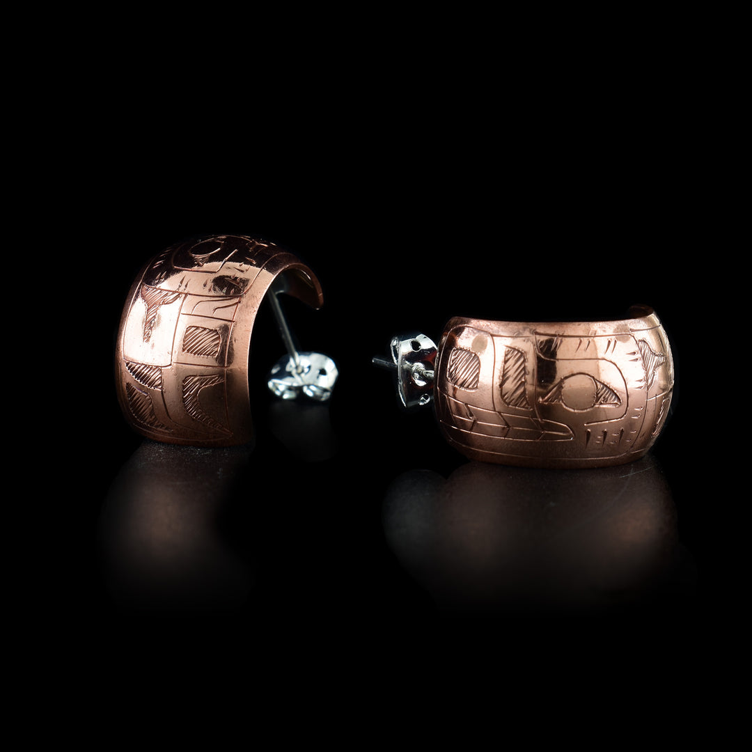 These copper hoop earrings have wide bands made from copper that are rounded and concaved. The bands for m small, half circles and there is a depiction of the Wolfs head carved into them. The nose, teeth, eyes and ears are shown and the head is facing the studs.