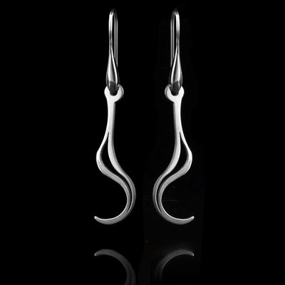 These sterling silver long dangling earrings have thin hangs that drop down and curve up at the ends. There is a cutout, teardrop shape in the center of them The hangs depict a Haida Gwaii character.
